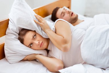 Horlama Konsepti. Kulaklarını yastıkla kaplayan, uyuyan kocanın yanında uyuyamayan, evde uyuyamayan sinirli kadın. Seçici Odaklanma