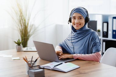 Müslüman Kadınlar İçin İş. Mutlu Arap kadın çağrı merkezi operatörünün portresi tesettür ve kulaklık takıyor, laptopta çalışıyor ve kameraya bakıyor.