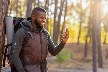 Sırt çantalı neşeli siyah adam akıllı telefonuyla sonbahar ormanlarının fotoğraflarını çekiyor.
