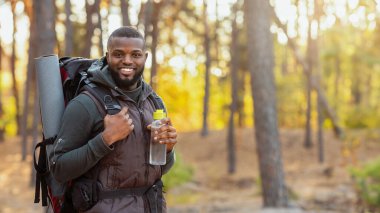Sırt çantalı gülümseyen Afrikalı genç adam sonbahar ormanı arka planında duruyor, elinde su şişesi, fotokopi alanı.
