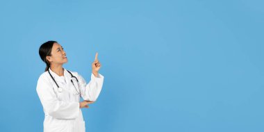 Boynunda steteskop olan Asyalı bir sağlık çalışanı beyaz önlük giymiş, koyu mavi bir arkaplanı olan düşünceli bir ifadeyle yukarıya doğru bakıyor.