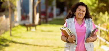 Neşeli bir öğrenci yeşil kampüs yolunda yürüyor, birkaç kitap taşıyor ve günlük giysiler giyiyor, çalışmalarını yaparken aydınlık ve güneşli bir günün tadını çıkarıyor..