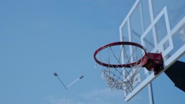 Güneşli bir öğleden sonra sergilenen parlak mavi gökyüzüne karşı duran bir basketbol potası. Çemberler rüzgarda hafifçe sallanıyor, oyuncuları basketbol oynamaya davet ediyor..