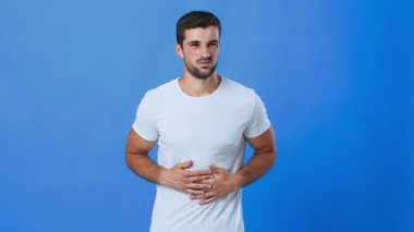 Genç bir adam mavi bir arka plana karşı duruyor, endişeli bir ifadeyle karnını nazikçe tutuyor, sağlığıyla ilgili rahatsızlık veya düşünce öneriyor..