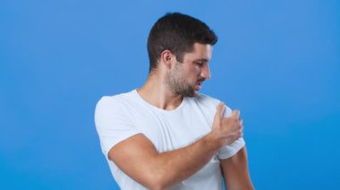 Düz beyaz t-shirt giyen genç bir adam hafifçe yana dönüyor, sol omzunu ovmak için eline uzanıyor, canlı mavi bir arka plana yaslanıyor..
