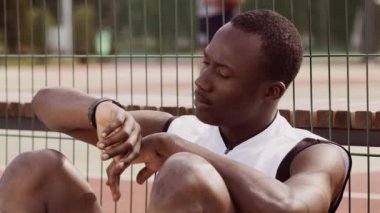 Afrika kökenli Amerikalı genç sporcu, yoğun bir antrenman sonrası saatini kontrol etmek için tenis kortunun kenarına oturur. Arka plan bulanık oyuncuların bir maçta yer aldığını gösteriyor.
