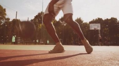 Genç bir siyah atlet, açık hava sahasında, parlak öğleden sonra güneşinin altında ustalıkla basketbol topu sürüyor. Diğer oyuncular arka planda antrenman yaparken görülebilir, canlı bir atmosfer yaratır..
