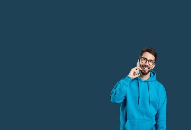 Mavi kapüşonlu bir adam arka planda duruyor, akıllı telefonuyla konuşurken gülümsüyor. Neşeli ve sohbete dalmış görünüyor, rahat bir tavır sergiliyor, boşluğu kopyalıyor.