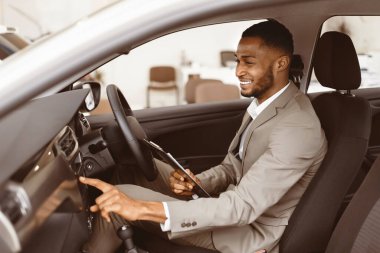 Car Insurance. Afro Agent Man Checking Vehicle Taking Notes Sitting In Automobile In Dealership Showroom. Selective Focus
