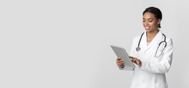 Çevrimiçi Doktor Danışmanlık. Gülümseyen siyah kadın terapist dijital tablet kullanıyor, steteskoplu beyaz önlüklü beyaz arka planda poz veriyor.
