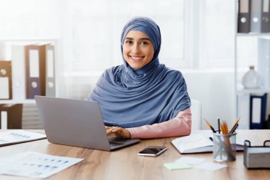 Müslüman Kadınlar İçin Modern İş. Ofiste dizüstü bilgisayarda çalışan mutlu İslamcı kız portresi ve kameraya bakmak, boşluğu kopyalamak