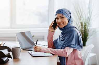 Modern ofiste dizüstü bilgisayarla çalışırken cep telefonuyla konuşan ve not alan Arapça kadın sekreterin portresi