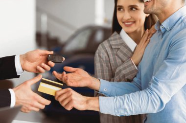 Buying Or Renting Car. Couple Giving Seller Credit Card And Taking Keys From Auto Standing In Dealership Office