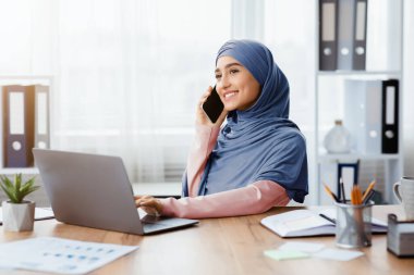 Gülümseyen Müslüman iş kadını cep telefonuyla konuşuyor, modern ofiste dizüstü bilgisayarda çalışıyor, İslami kadın çalışan telefonla masada oturuyor.