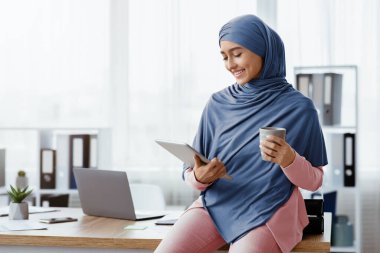 İşteyken gevşe. Genç Suudi iş kadını tesettürde kahve içiyor ve dijital tablet kullanıyor, ofiste dinleniyor, boş yer arıyor.