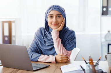 Müslüman kadın girişimci modern ofiste masada oturuyor ve kameraya poz veriyor, işyerinde dinleniyor, boş alan
