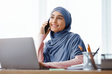 İş kariyeri. Kendine güvenen Arap iş kadını, modern ofiste dizüstü bilgisayar kullanırken cep telefonuyla konuşuyor.