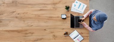 İş İletişimi. Modern ofiste masa başında otururken akıllı telefon kullanan ve dizüstü bilgisayarda çalışan Arapça kadın çalışanın en iyi görünümü