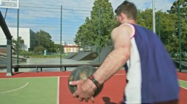 Kolsuz tişörtlü bir adam basketbola doğru koşuyor. Atışa hazır. Arka planda kaykay rampaları var..