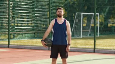 Güneşli bir günde, bir adam basketbol sahasında, spor kıyafetlerini giymiş bir şekilde elinde bir top tutuyor..