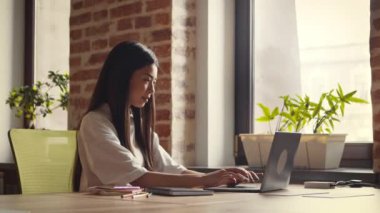 Asyalı kız iyi ışıklandırılmış, davetkar bir çalışma alanındaki bitkilerle çevrili bir masada otururken dizüstü bilgisayarına odaklanıyor..