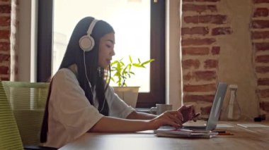 Asyalı bir kız, ahşap bir masada oturuyor, kulaklık takarken dizüstü bilgisayarına odaklanıyor, arka planda bir bitki ve pencereden gelen doğal ışık var..