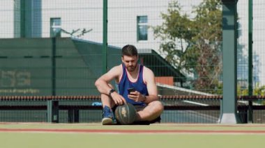 Atletizm kıyafetleri içindeki genç bir adam basketbol sahasında oturuyor, telefonuna odaklanmış, yanında bir basketbol topuyla, güpegündüz..