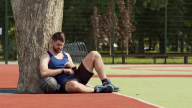 Genç bir adam basketbol sahasında bir ağaca yaslanır, topu tutar ve gevşemek için zaman ayırır..