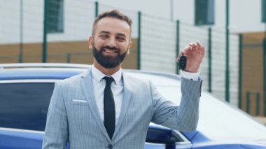 Bir adam arabanın anahtarlarını tutarken gülümsüyor, açık gökyüzünün altında modern bir kentsel ortamda mavi bir aracın yanında duruyor..