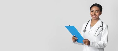 Beyaz önlüklü genç afro kadın doktor elinde mavi bir pano ve ellerinde boş bir arkaplanda duran bir hastanın klinik hikayesi var.