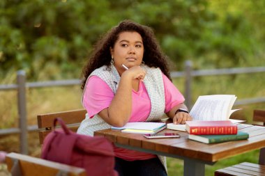 Bir öğrenci açık bir masada oturuyor, etrafı kitaplar ve defterlerle çevrili. Düşünceli görünüyor, parlak bir günde yeşilliğin tadını çıkarırken notlar yazıyor..