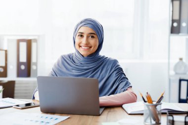 Female Entrepreneurship. Portrait of happy arabic businesswoman in hijab using laptop at workplace and smiling at camera, empty space