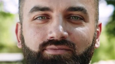 Sakallı ve yeşil gözlü bir adam açık hava ortamında sıcacık gülümsüyor..
