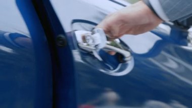 Bir el, gündüz vakti şehrin aydınlık bir bölgesinde park etmiş mavi bir arabanın krom sapını tutuyor..