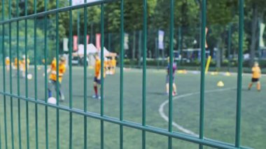 Turuncu formalı genç oyuncular ağaçlar ve sancaklarla çevrili yeşil bir alanda futbol antrenmanı yapıyorlar..