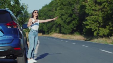 Mavi bir arabanın yanında duran bir kadın, güneşli bir günde sakin bir köy yolunda yol alabilmek için kolunu uzatıyor..