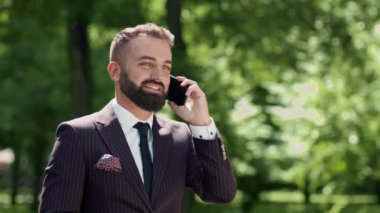Çizgili takım elbiseli bir adam, güneş ışığıyla aydınlatılmış yemyeşil bir parkta dikilirken cep telefonuyla konuşuyor..