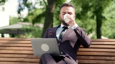 Takım elbiseli bir adam dizüstü bilgisayarını kullanır ve parktaki bir bankta kahvesini yudumlar. Etrafı yeşilliklerle çevrilidir ve güneşli havanın tadını çıkarırlar..