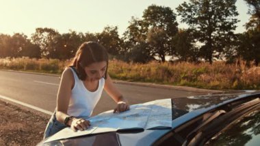 Bir kadın gün batımında sakin bir manzarayla çevriliyken, bir arabanın kaputunda bir haritanın üzerine eğilir ve rotasına odaklanır..