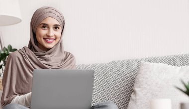 Serbest meslek kavramı. Türbanlı mutlu milenyum kızı kucağında dizüstü bilgisayarla kanepeye oturuyor ve kameraya bakıyor.