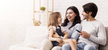 Happy Parenthood. Young Parents Enjoying Spending Time With Their Little Daughter At Home, Watching Tv In Living Room Together
