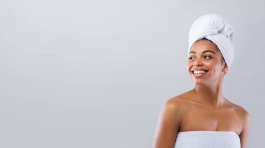 Happy young african woman wrapped in white bath towels after shower looking at empty space, grey studio background, discounts at spa hotel