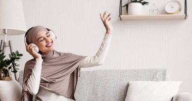 Music streaming services concept. Joyful muslim girl in headphones enjoying favorite melody and rhythm sitting on comfy couch at home