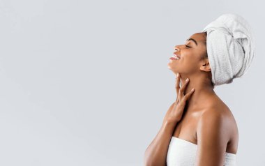 Duş aldıktan sonra beyaz banyo havlularına sarılmış neşeli Afrikalı genç bir kadının yan görüntüsü, ipeksi teninin tadını çıkarması, boş bir yer, gri stüdyo arka planı, spa veya saunada indirimler.