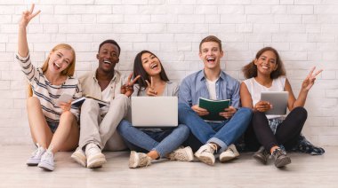 Üniversite sınavlarına çalışan bir grup mutlu çok ırklı öğrenci, kameraya neşeli bir şekilde el kol hareketi, beyaz tuğla duvar arka planı.
