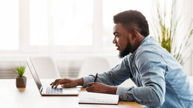 Genç Afro-Amerikan çalışan dizüstü bilgisayar kullanarak ve modern ofiste çalışırken notlar alarak, kopyalama alanı ile yan görünüm