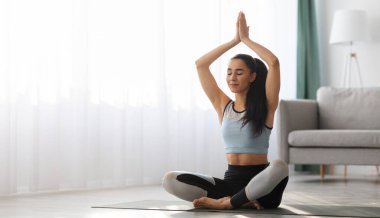 Spor kıyafetli genç bir kadın evde yoga yapıyor, atletik esmer bayan yoga minderinde gözleri kapalı oturuyor ve oturma odasında meditasyon yapıyor, fotokopi çekiyor. Yoga, sağlıklı yaşam tarzı konsepti