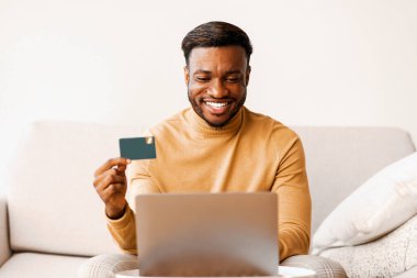 İnternetten alışveriş. Mutlu Afrikalı Amerikalı adam kredi kartını evdeki koltukta otururken dizüstü bilgisayar kullanarak satın alıyor..