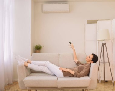 Akıllı Ev. Mutlu kız uzaktan kumandayı tutuyor klimanın altında dinleniyor, kanepede uzanıyor, kopyasal alan