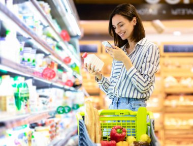 Gülümseyen genç bir kadın markette raftan süt ürünleri alıyor, elinde şişe ve akıllı telefon, cep telefonuyla barkodları tarıyor, el arabası ile yürüyor.